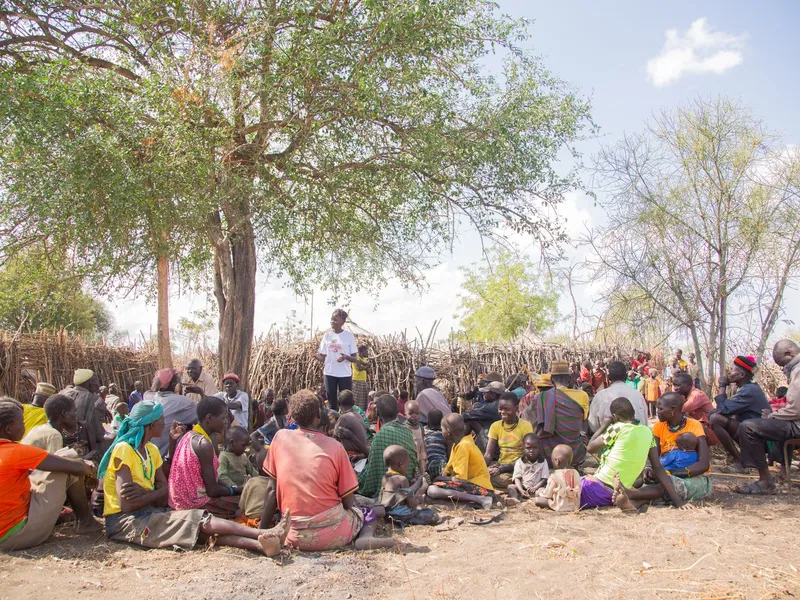 Adults and children gather in a large circle under a tree for an outdoor community meeting