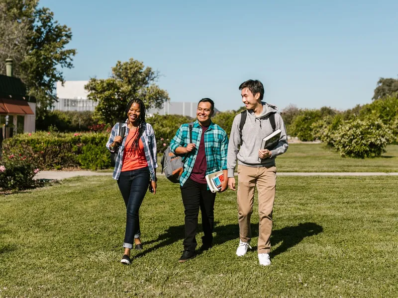 Three college students with backpacks and textbooks walking together across a sunny green campus lawn