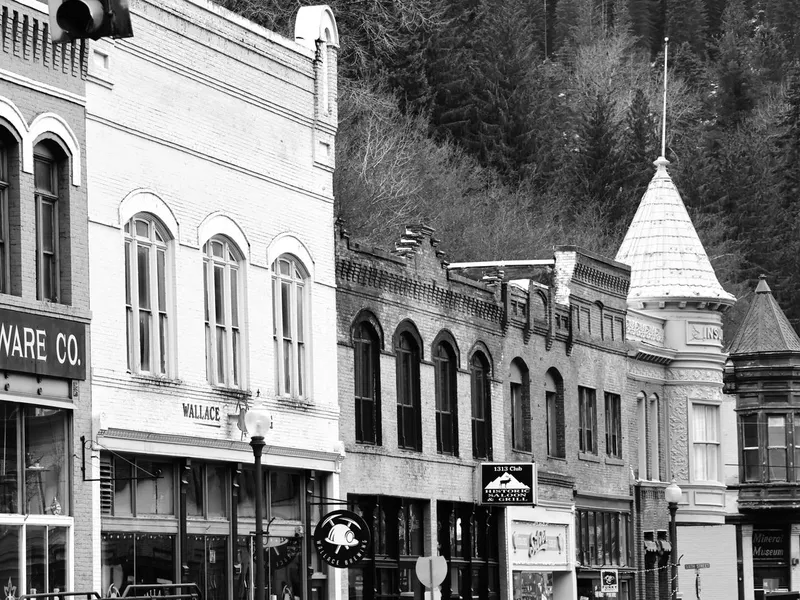 Historic brick mining-era storefronts with turrets along a street in downtown Wallace, Idaho, below a wooded hillside
