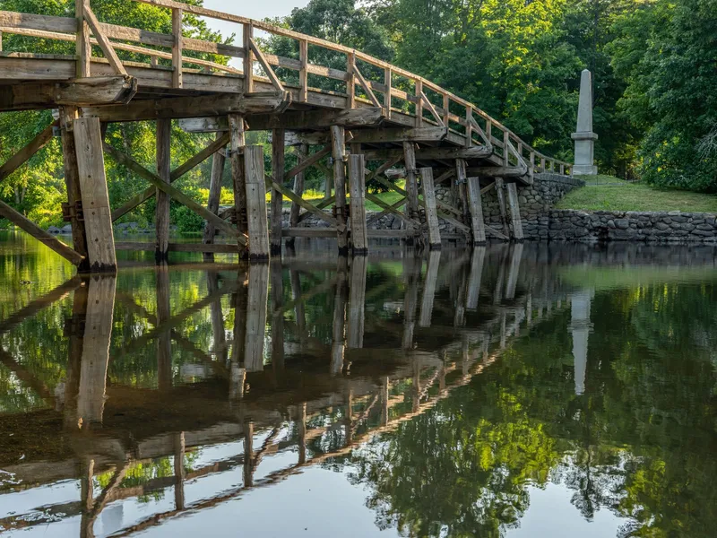 Old North Bridge in Concord, Massachusetts, reflected in calm water with a stone monument among trees