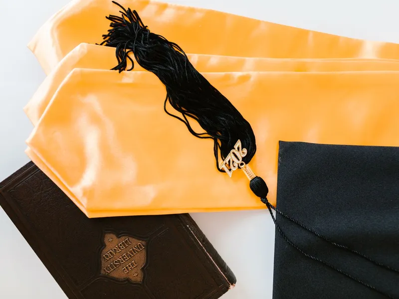 Graduation cap, tassel and gold stole resting beside a leather-bound book on a white table