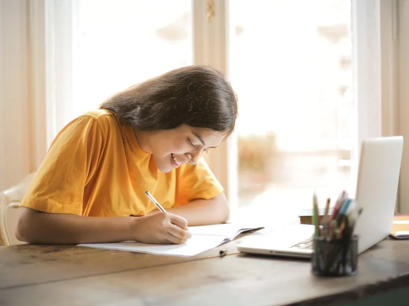 Young woman studying at home with a laptop and notepad, smiling while she writes