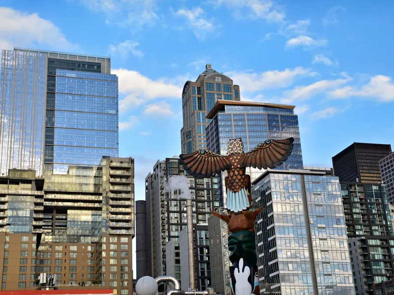 Carved and painted eagle totem pole rising in front of Seattle downtown towers under a blue sky
