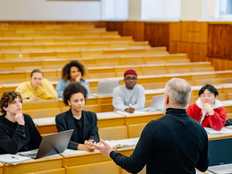Professor addressing economics students seated with laptops at long wooden desks in a university lecture hall