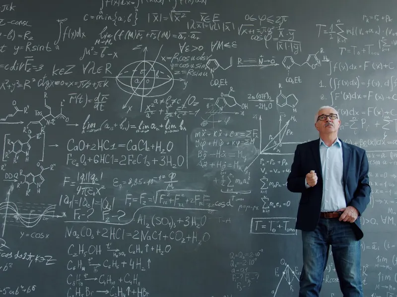 Professor in a blazer standing before a blackboard covered in dense equations and formulas