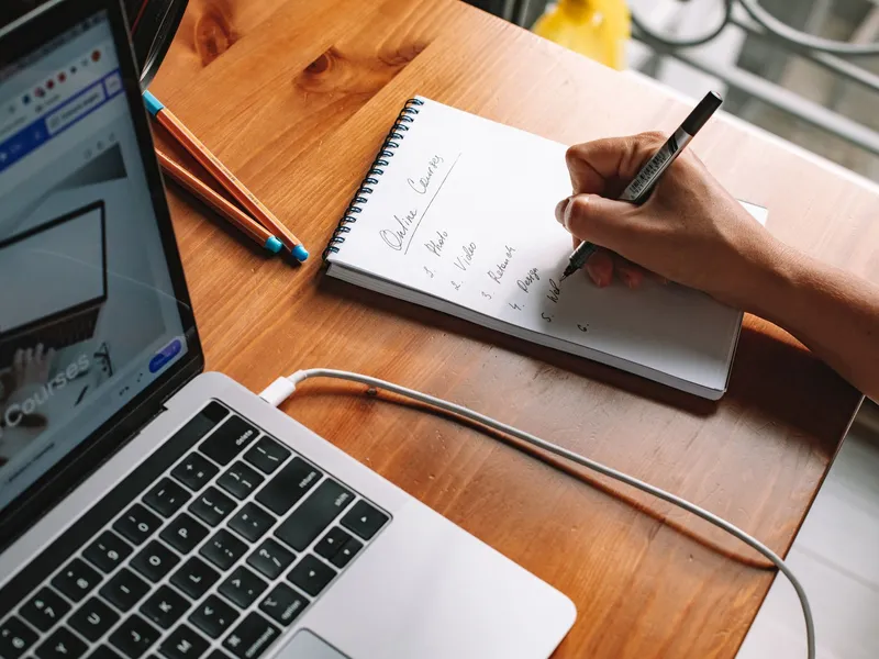 Student taking handwritten notes beside a laptop showing an online course