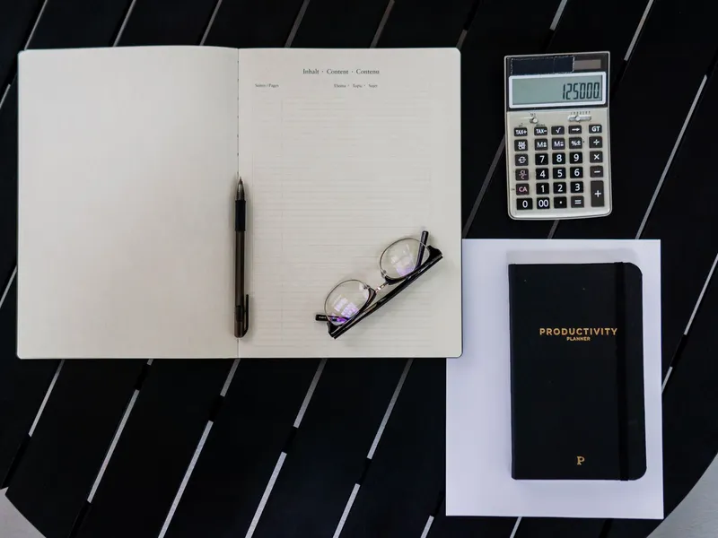 Open notebook, pen, reading glasses, calculator and planner laid out on a black table for budget planning
