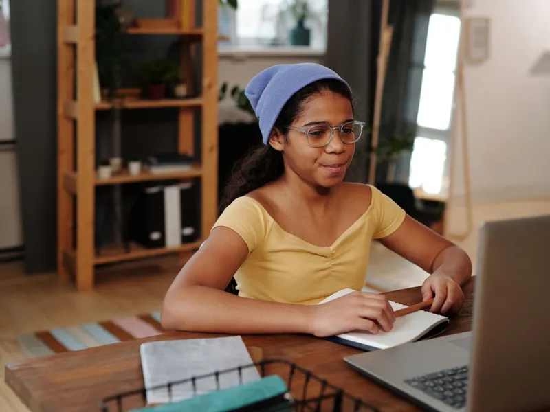 Girl in a blue beanie studying remotely on a laptop at a table at home