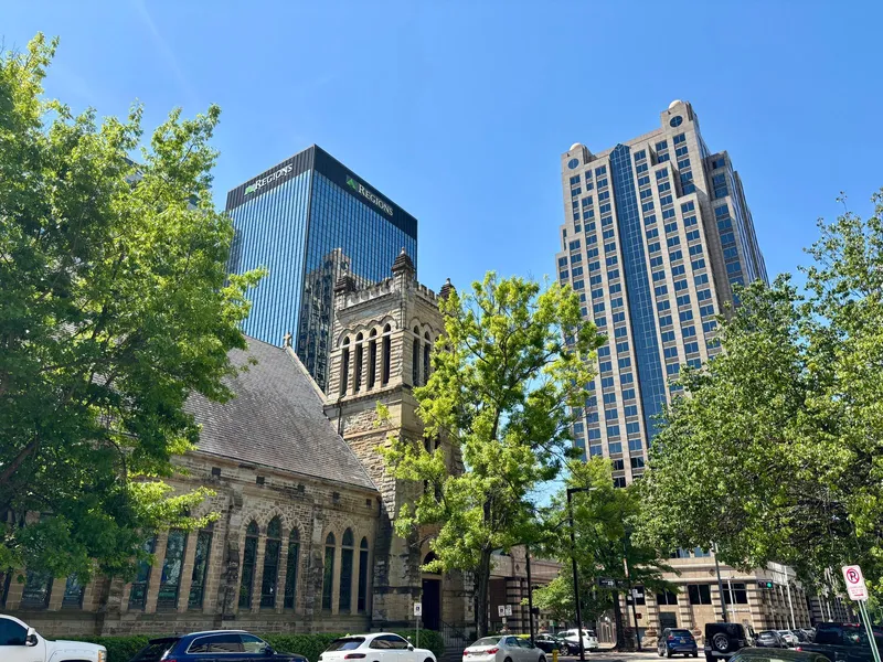 Historic stone church standing beside glass skyscrapers on a tree-lined downtown street in Birmingham, Alabama