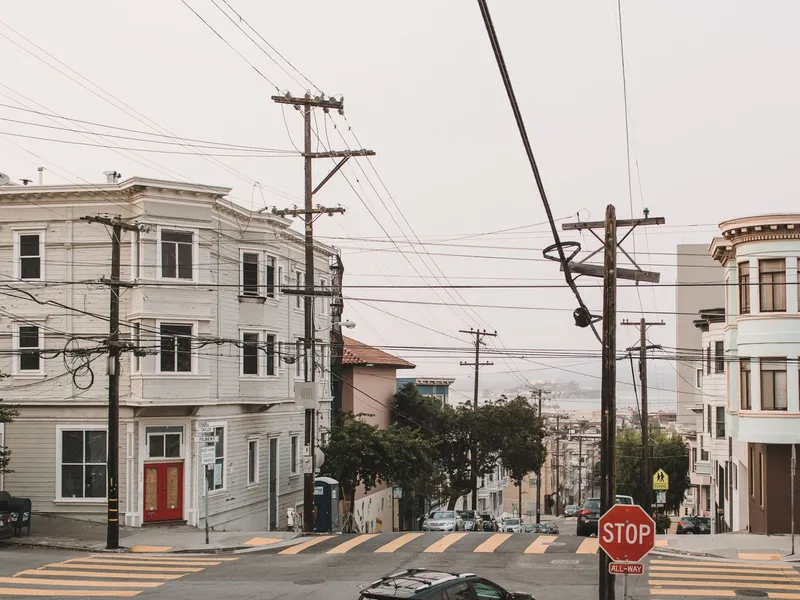 Steep San Francisco intersection with Victorian apartment buildings, utility poles and a distant view toward the bay