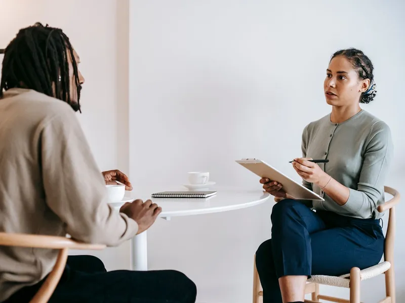 Interviewer taking notes on a clipboard while talking with a participant at a small table