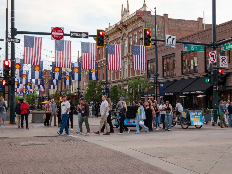 Pedestrians crossing a downtown Denver intersection lined with brick buildings and hanging American and Colorado flags