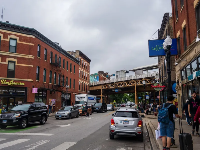 Busy Chicago neighborhood street with brick buildings, shops, pedestrians and an elevated train line overhead