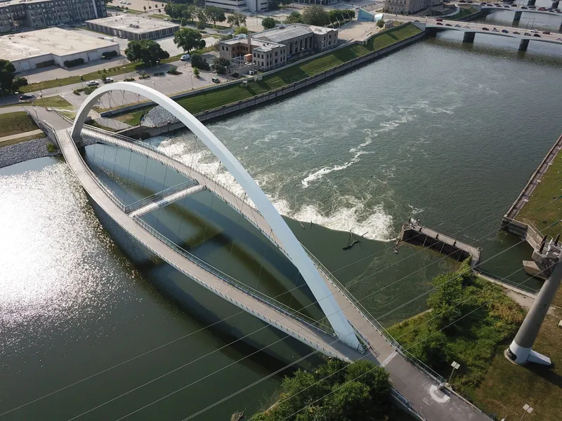 Aerial view of a white arched pedestrian bridge over the river beside downtown buildings and roads in Des Moines, Iowa