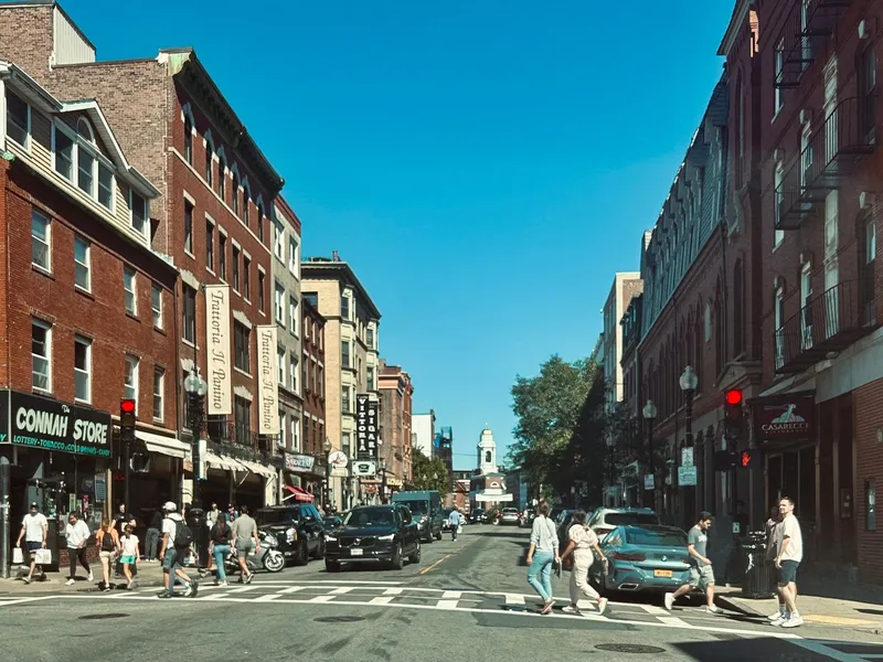 Pedestrians crossing Hanover Street in Boston's North End lined with red brick buildings and shops