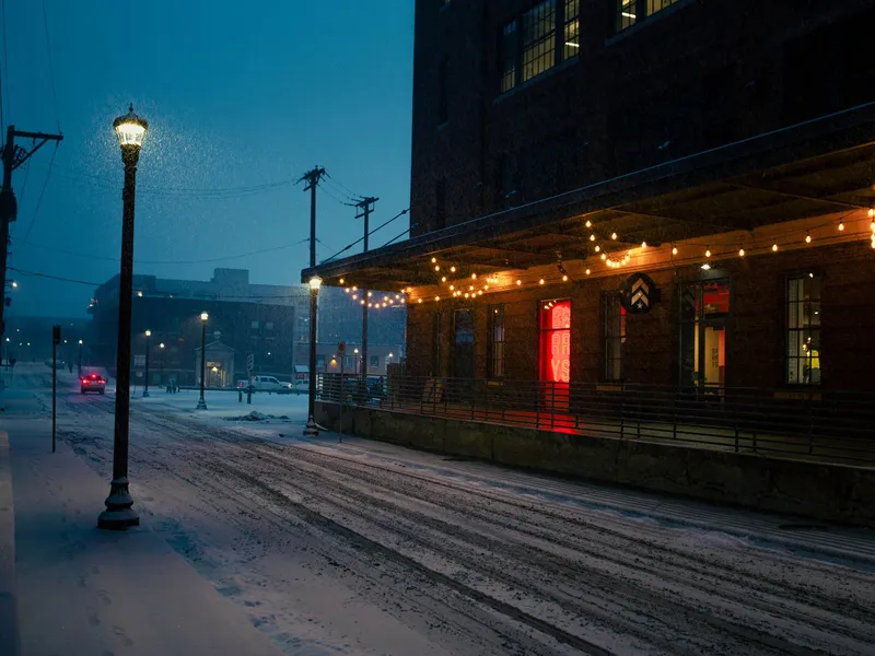 Snowy Minneapolis street at dusk with a glowing streetlamp and string lights on a brick building