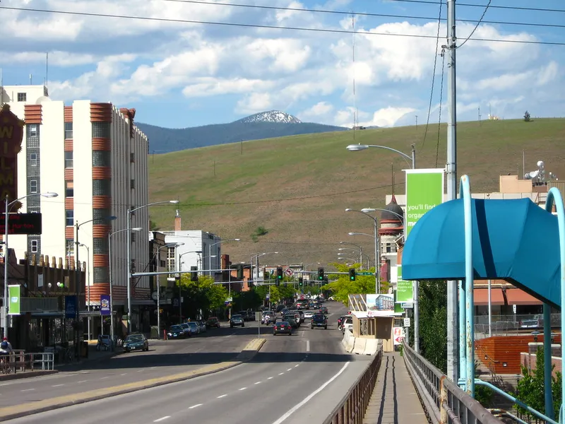 Downtown Missoula street with mid-rise buildings, traffic and a green mountain hillside in the distance