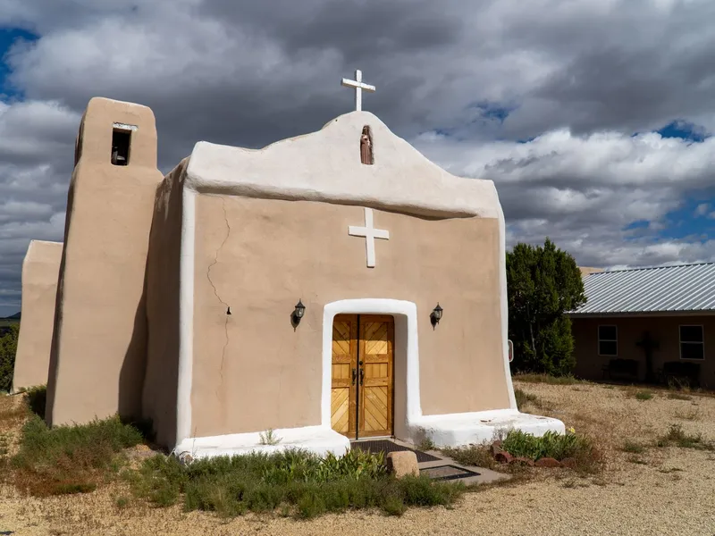 Small adobe church with a bell tower and wooden doors in a New Mexico village under a cloudy sky