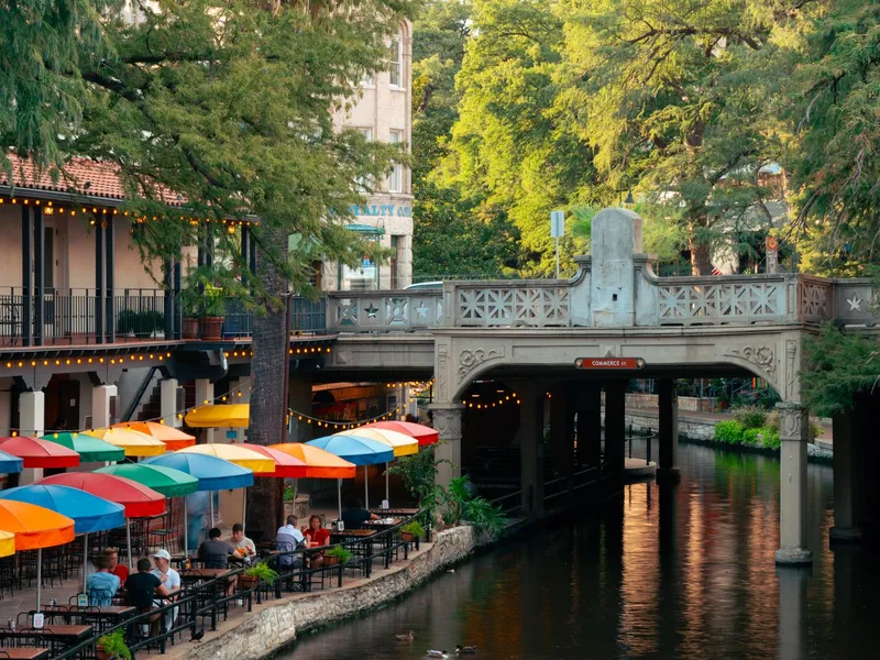 Diners under colorful umbrellas beside the San Antonio River Walk canal and a stone bridge