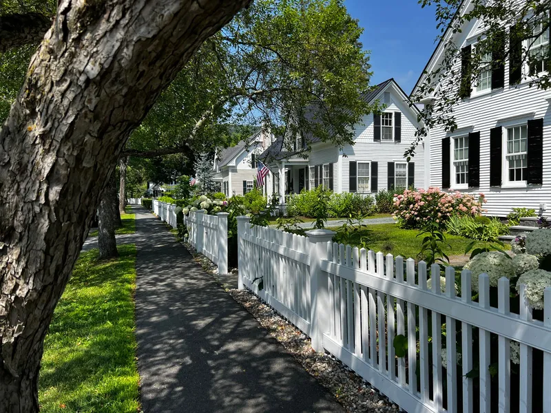 Sunlit village sidewalk lined with white picket fences and clapboard houses in New England