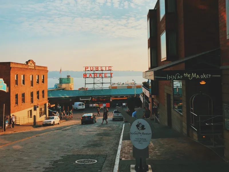 Pedestrians cross a brick street in Seattle toward the Public Market sign and Elliott Bay at sunset