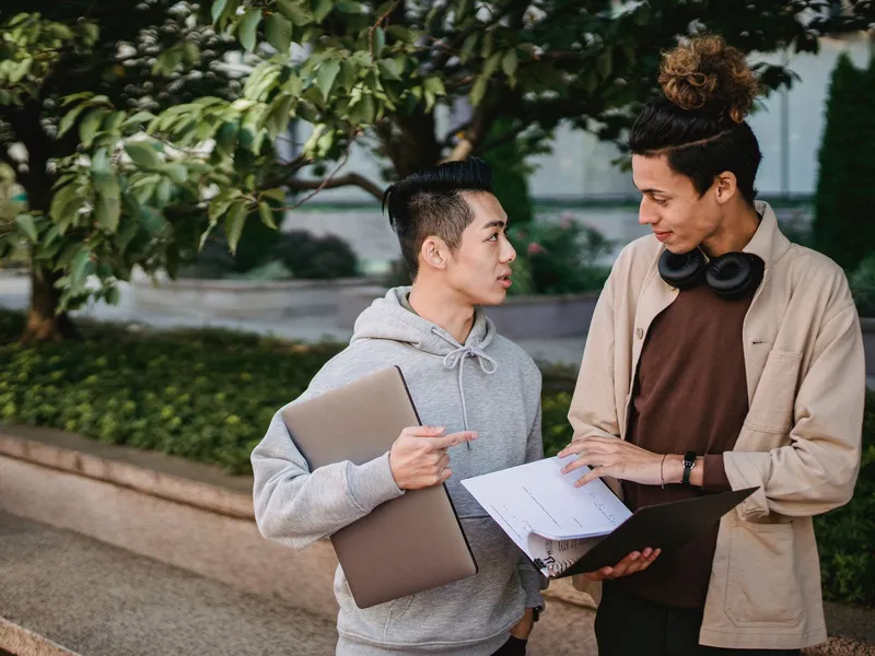 Two students with textbooks and a folder discussing coursework while standing in a campus park