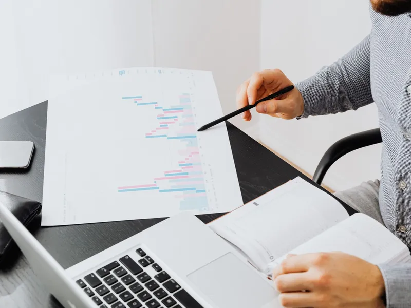 Person pointing at a graph printout beside an open laptop and notebook during data analysis