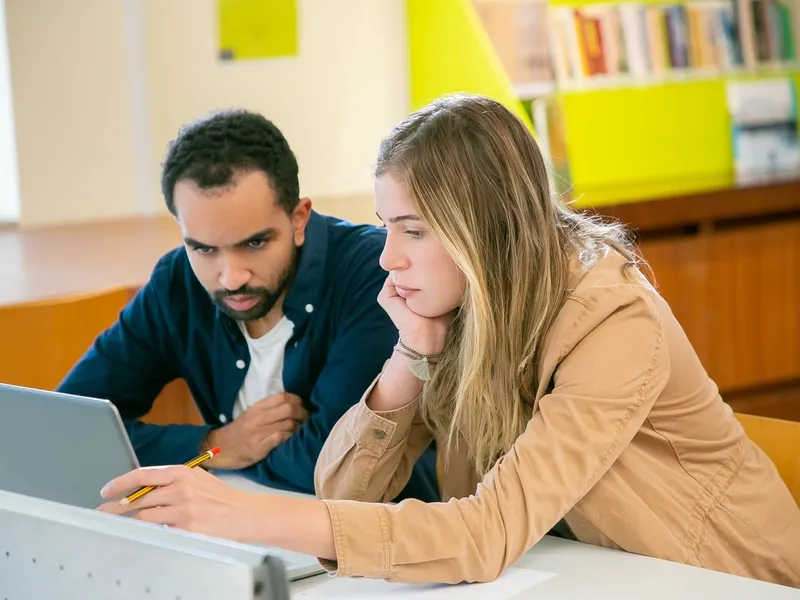 Two classmates working together on a laptop with books in a university library