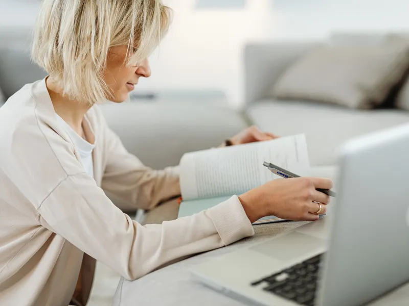 Woman with a book and laptop on a sofa researching online degree options