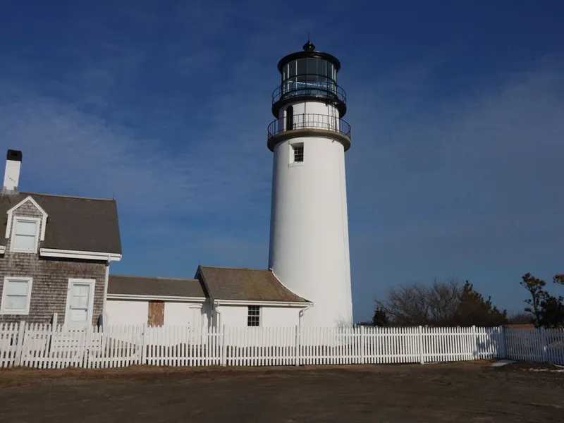 White Cape Cod lighthouse beside a shingled keeper's house and picket fence under a deep blue sky in Truro