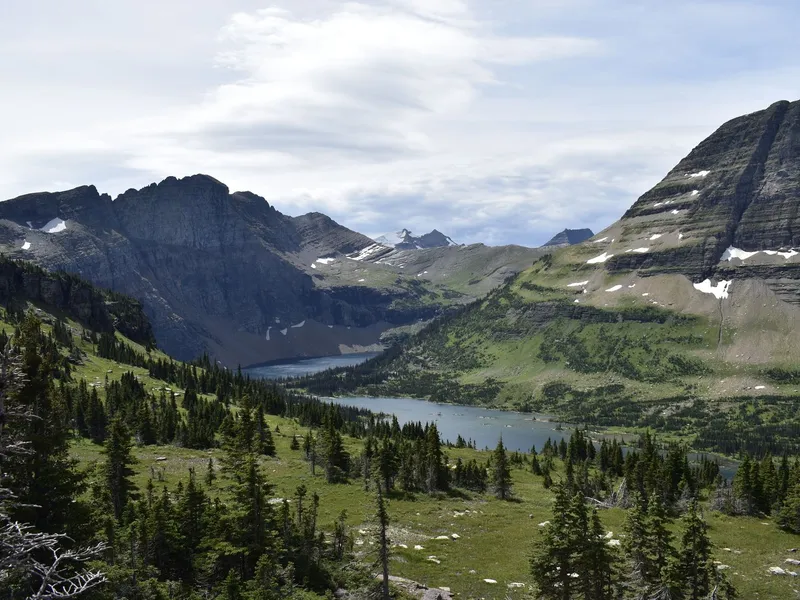 Mountain valley with a lake, pine forest and rocky peaks with patches of snow under a cloudy sky