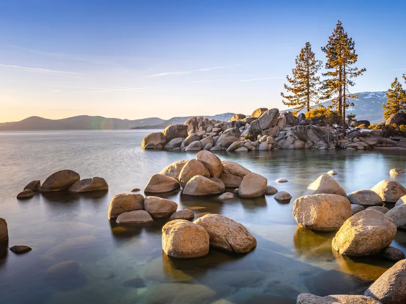 Granite boulders and pine trees along a clear mountain lake shoreline at sunset